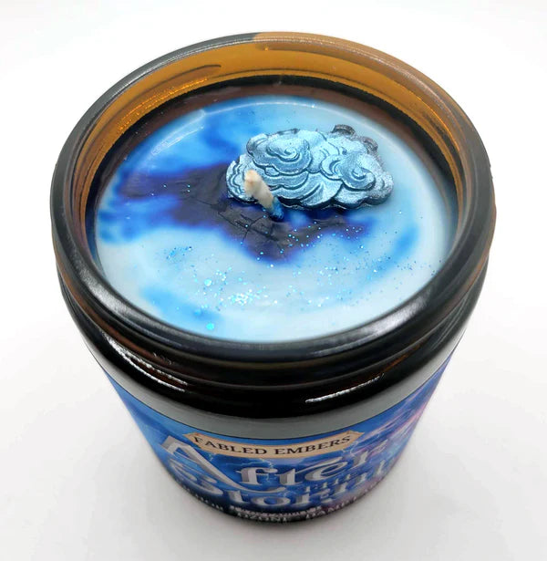A glass jar with a candle inside, the top of the candle is a swirl of light blue and dark blue wax, with silver glitter sprinkled in places. There is a small blue and white cloud made of wax inlaid on top.